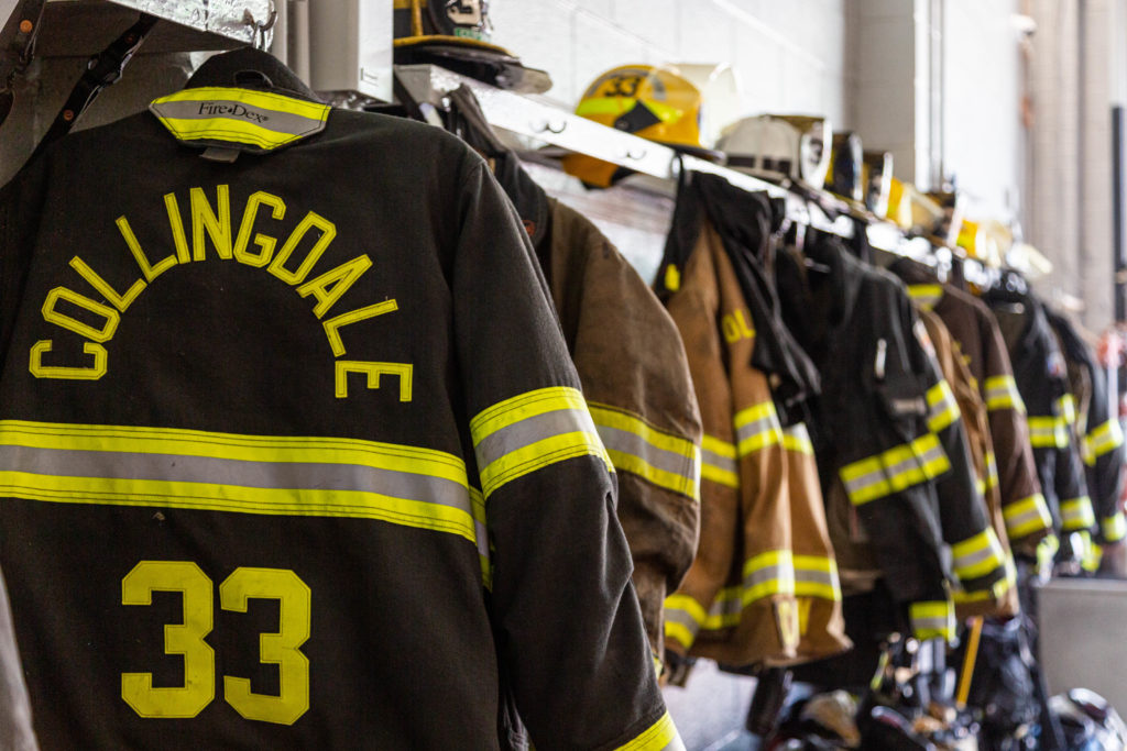 A row of firefighter jackets and helmets hanging on hooks in a fire station, with the closest jacket labeled "COLLINGDALE 33.
