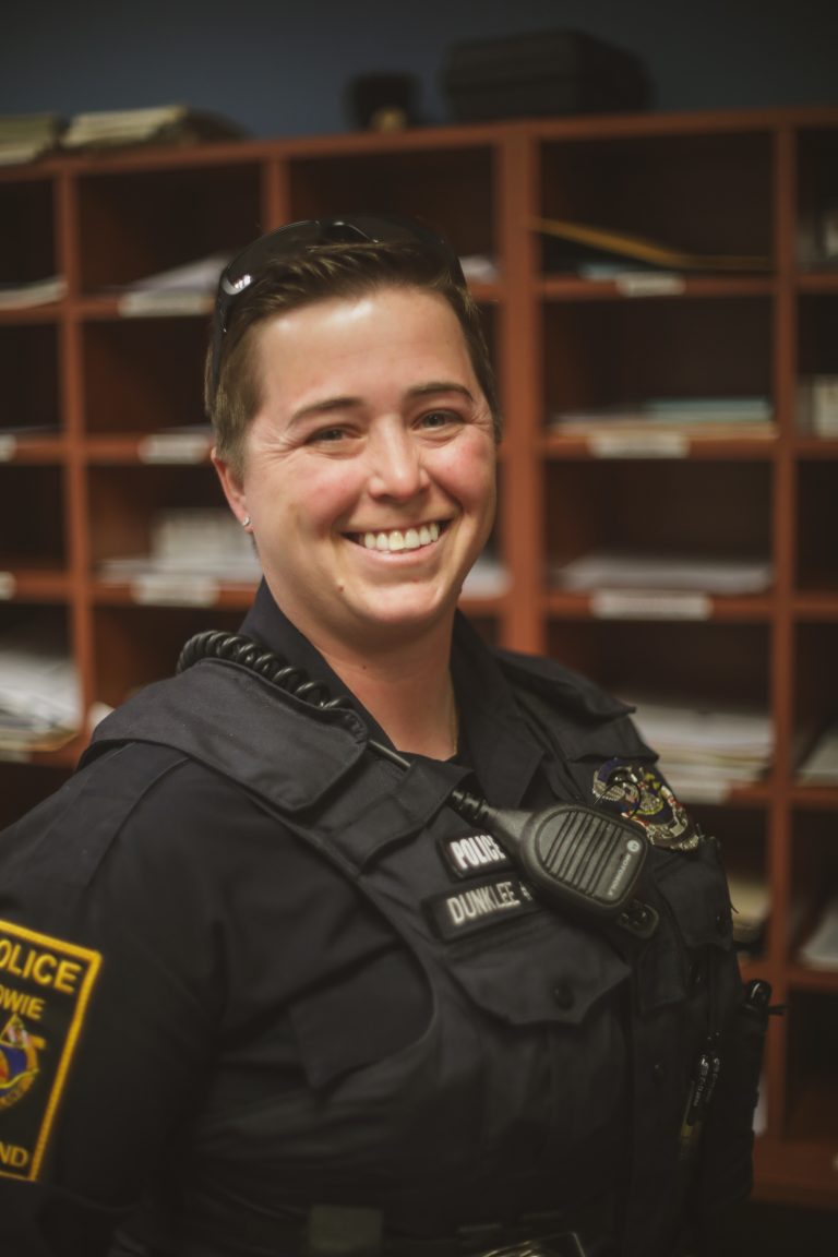 A smiling police officer stands indoors, wearing a uniform and a vest with a badge and radio. Behind them are shelves filled with files and papers.