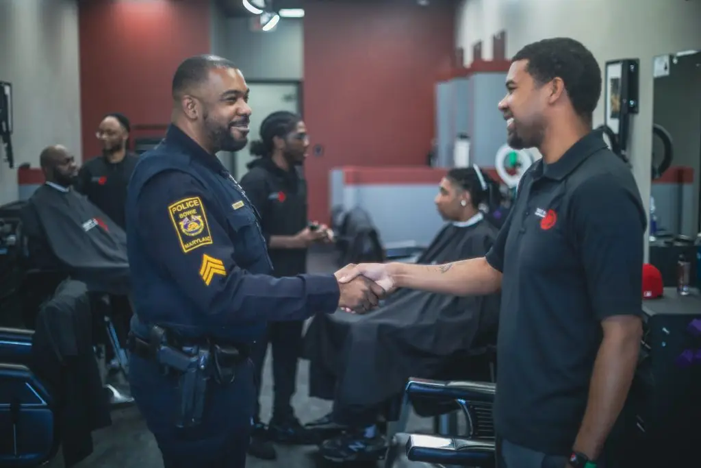 A police officer in uniform and a man in a black polo shirt shake hands in a barbershop, with other men sitting in barber chairs and barbers working in the background.