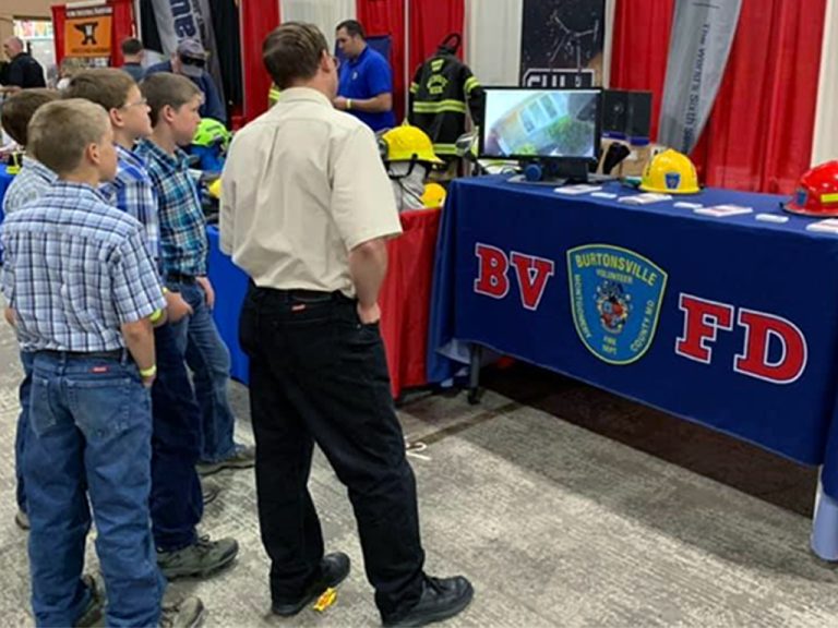 A group of boys stands in front of a Burtonsville Volunteer Fire Department table at an indoor event, watching a monitor. The table displays the BVFD logo, helmets, and fire safety materials.