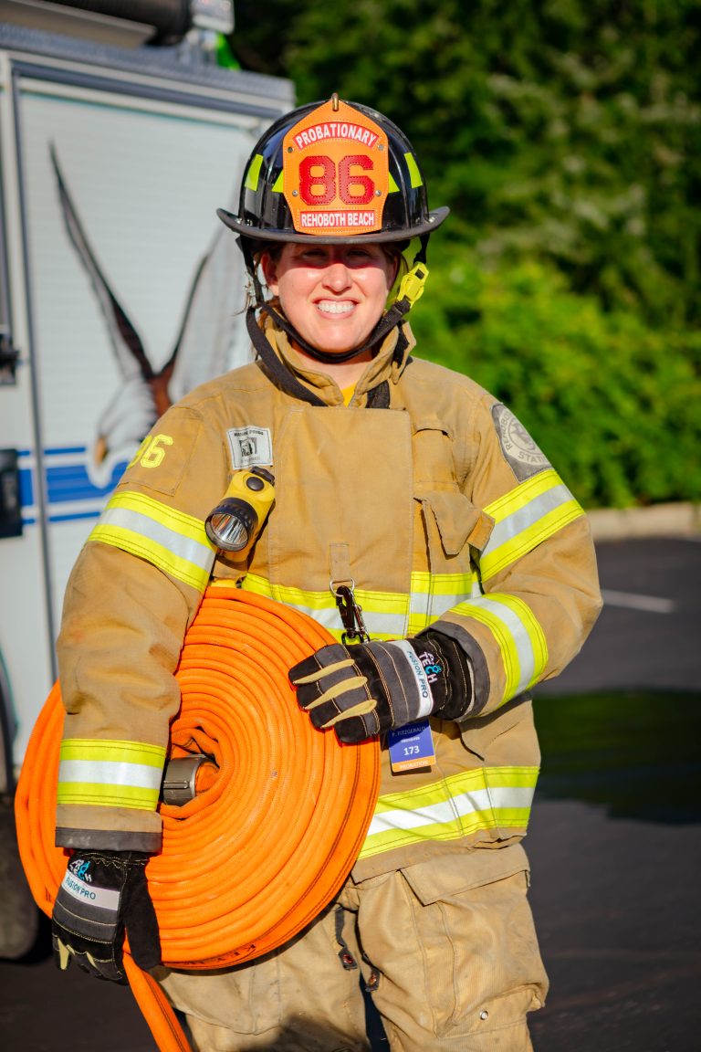 A firefighter in full gear, wearing a helmet labeled "Probationary 86 Rehoboth Beach," stands outdoors holding an orange fire hose and smiling, with a fire truck and trees in the background.