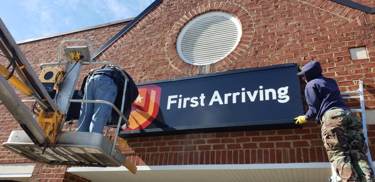 Two workers are installing a black sign with the text "First Arriving" and a shield logo on the brick exterior of a building, using a lift and wearing work clothes and gloves.