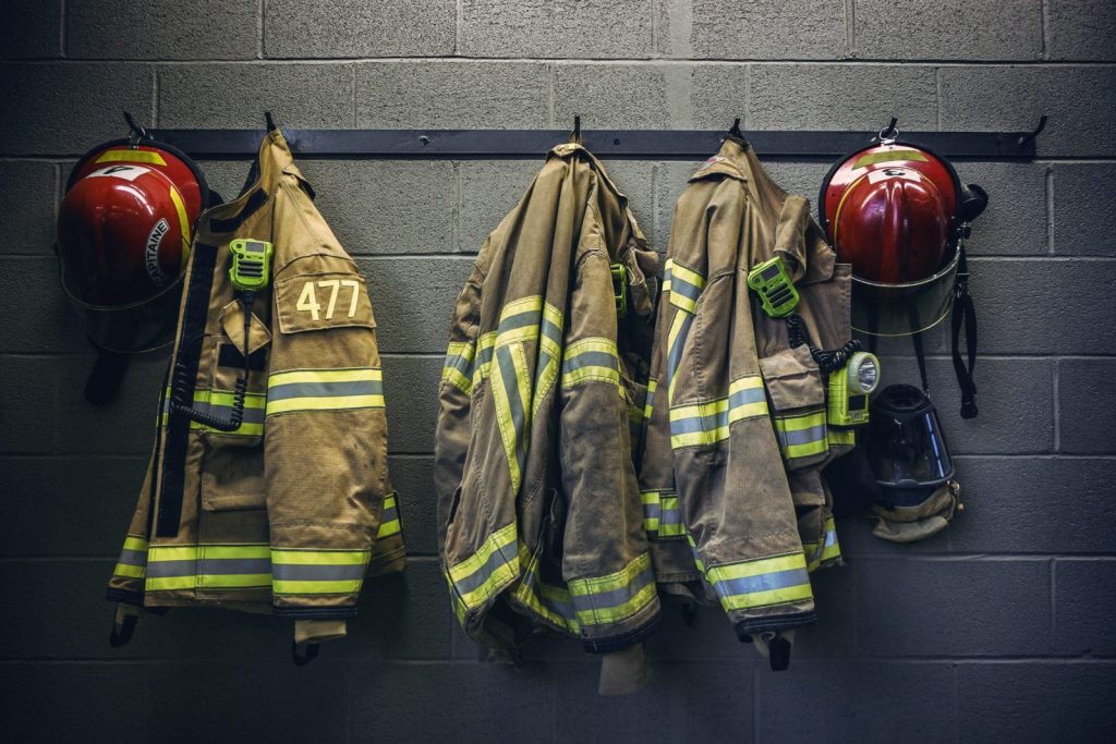 Three firefighter jackets with reflective stripes are hanging on hooks, each with a radio clipped to it. Two red helmets and a breathing mask are also hanging on a gray cinder block wall.