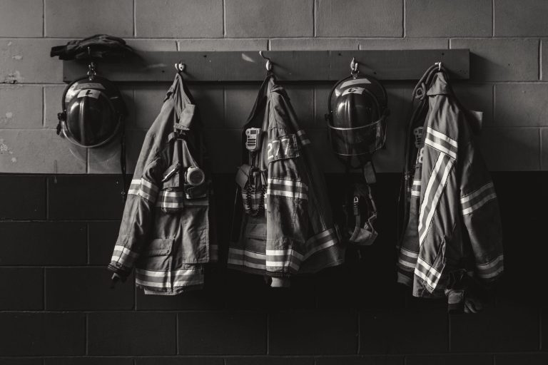 Black and white photo of four firefighter uniforms, including helmets and jackets with reflective stripes, hanging on hooks against a brick wall. One set has a pair of gloves on top of the helmet.