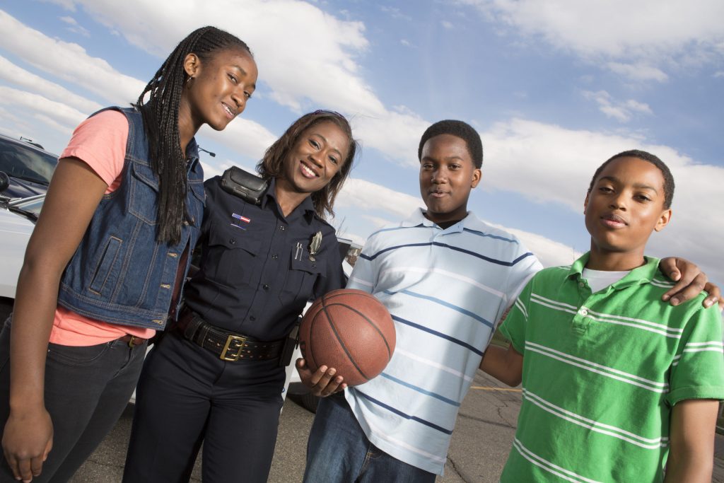 A smiling police officer stands outdoors with three teenagers, one holding a basketball. The group is posing together, showing friendliness and community engagement, with a police car and blue sky in the background.