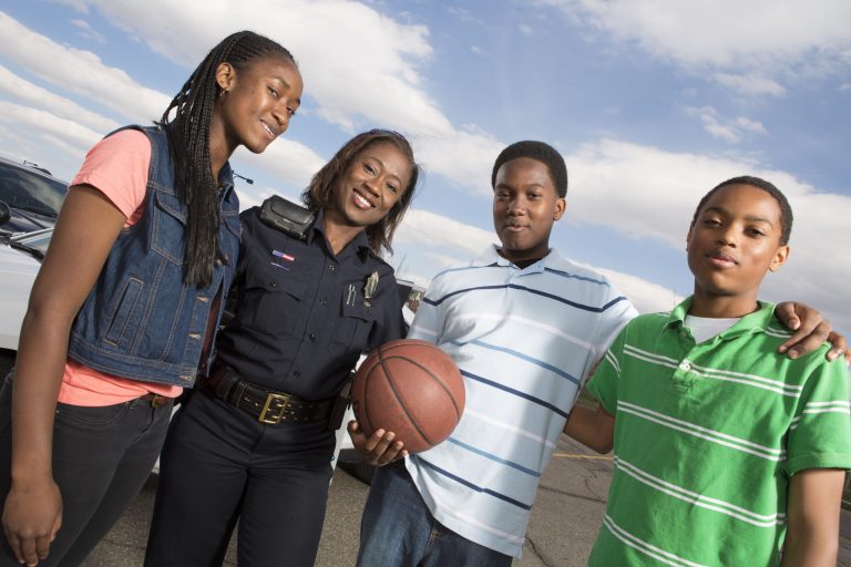 A smiling police officer stands outdoors with three teenagers, one holding a basketball. The group is posing together, showing friendliness and community engagement, with a police car and blue sky in the background.