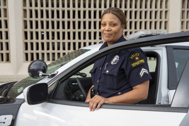 A police officer in uniform stands beside an open car door, resting one hand on the vehicle and smiling at the camera. A building with a grid-like facade is visible in the background.