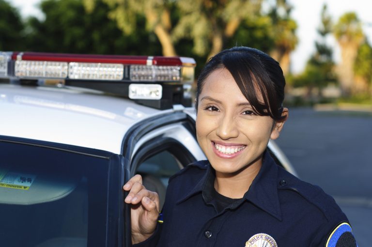 A smiling police officer stands beside an open car door, wearing a dark uniform with a badge, in front of a police car, with trees and greenery visible in the background.