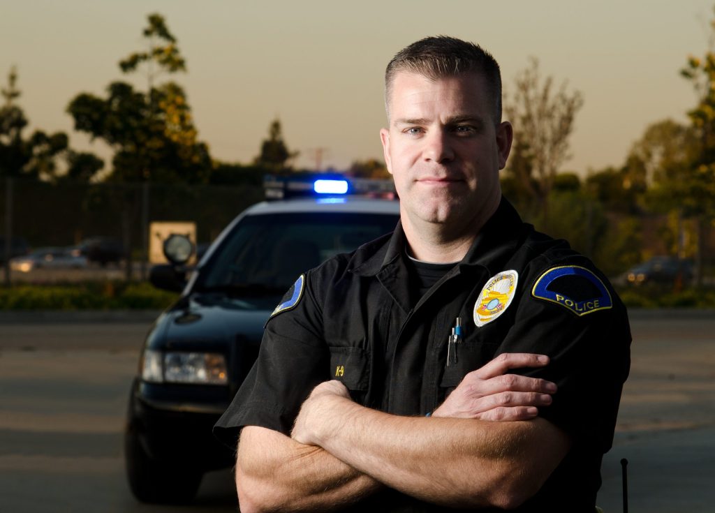 A police officer stands with arms crossed in front of a patrol car with flashing lights, outdoors in a parking lot at dusk. Trees and a fence are visible in the background.