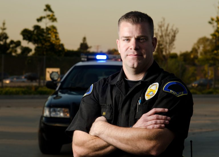 A police officer stands with arms crossed in front of a patrol car with flashing lights, outdoors in a parking lot at dusk. Trees and a fence are visible in the background.