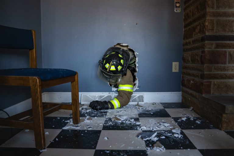 A firefighter in full gear crawls through a hole in a wall, surrounded by debris, in a room with checkered tile flooring, a chair, and a brick fireplace.