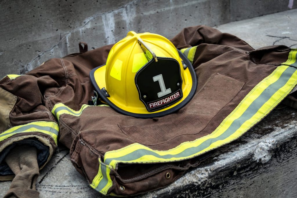 A yellow firefighter helmet labeled “1 Firefighter” rests on a brown fire jacket with yellow reflective stripes, both placed on a concrete surface.