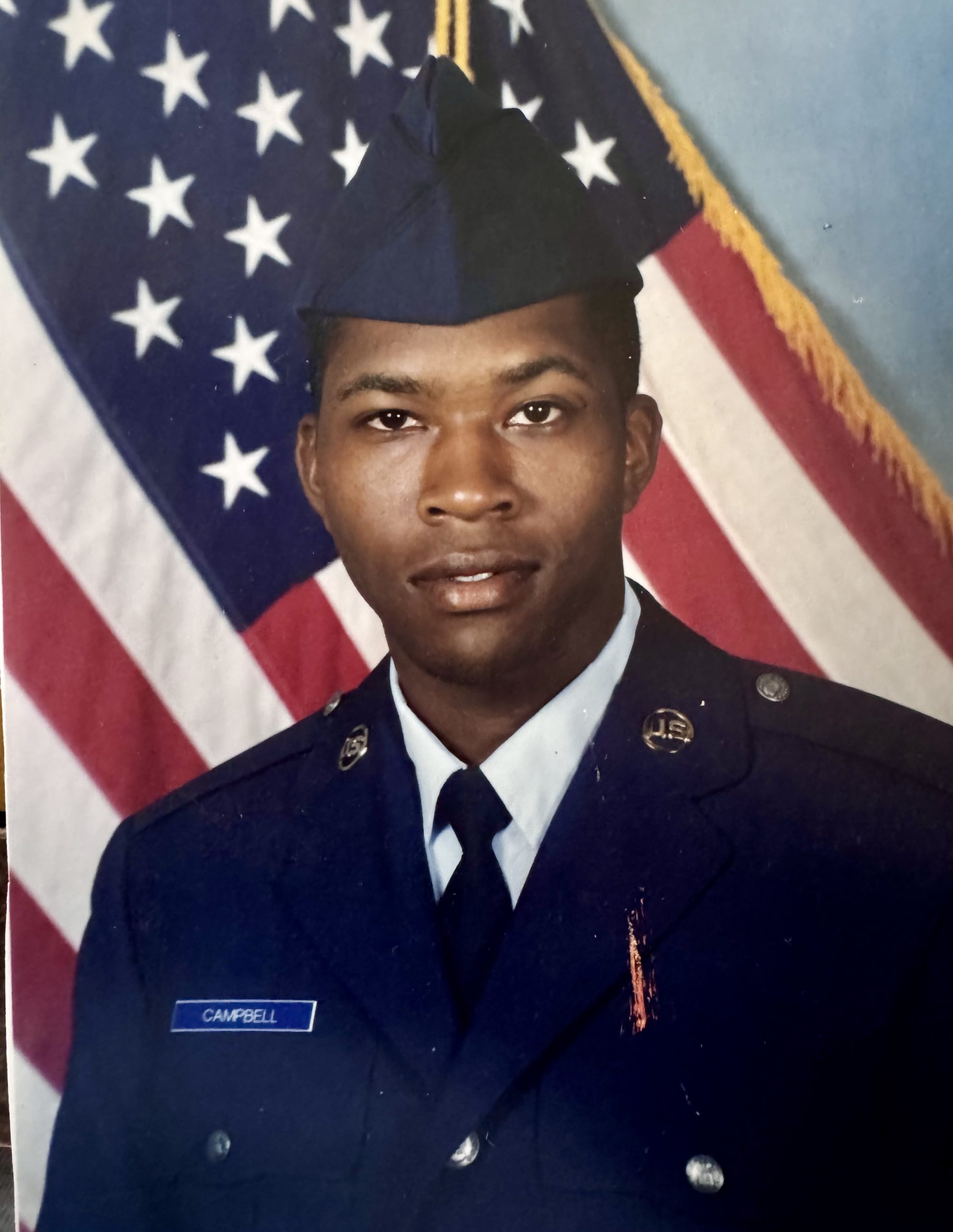 A man in a formal U.S. Air Force uniform poses for a portrait in front of an American flag, wearing a blue cap and a name tag that reads "Campbell.