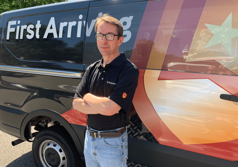 A man wearing glasses and a black polo shirt stands with arms crossed in front of a van with the words "First Arriving" and a large star graphic on its side.