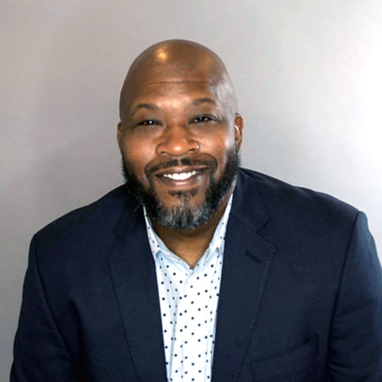 A man with a bald head and short beard smiles, wearing a navy blazer over a white shirt with small black dots, posed against a plain light gray background.