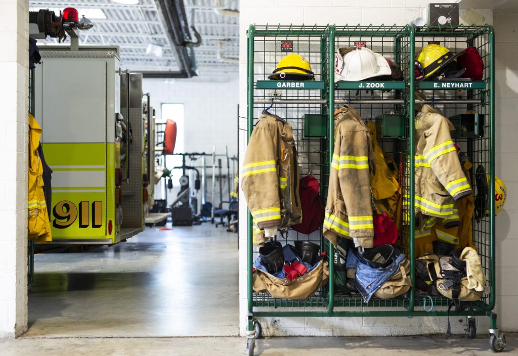 Three firefighter lockers holding helmets, jackets, and boots are seen inside a fire station. A fire engine with the number 911 is parked nearby, and workout equipment is visible in the background.