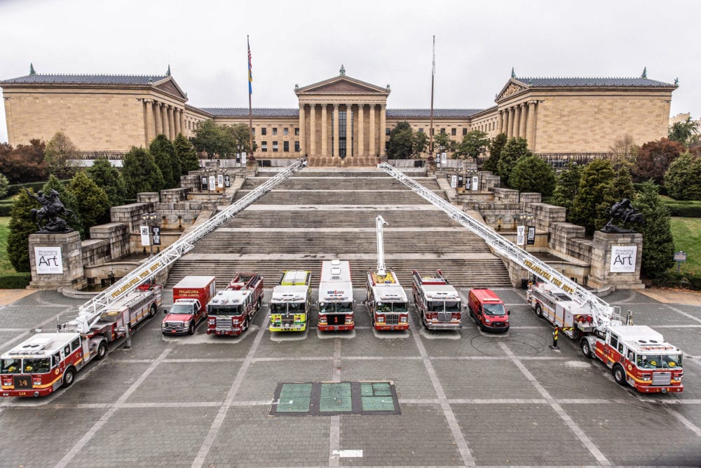 Several fire trucks and emergency vehicles are parked in front of the Philadelphia Museum of Art, lined up at the base of the famous steps on a cloudy day.