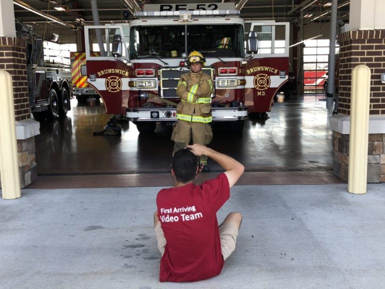 A person in a red "First Arriving Video Team" shirt sits on the ground, taking a photo of a firefighter posing in front of a red Brunswick fire truck inside a fire station garage.