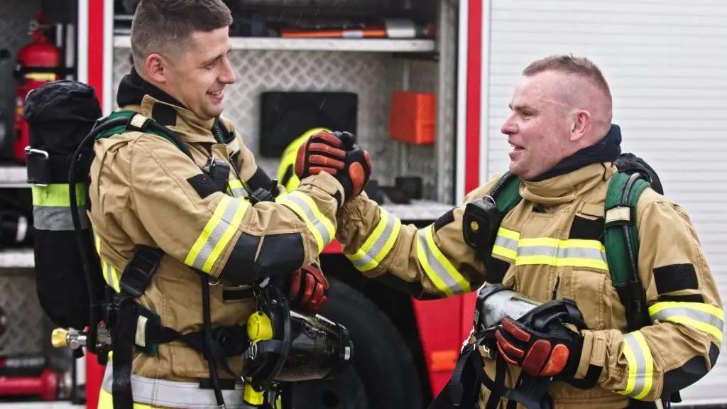 Two firefighters in tan protective gear and reflective stripes smile and shake hands in front of a fire truck, showing camaraderie and teamwork. Both wear gloves and carry equipment.