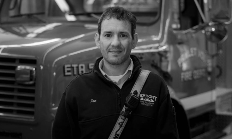 A man in a uniform stands in front of a fire truck. He wears a jacket with a name badge that says "Vermont Fire Academy." The photo is in black and white.