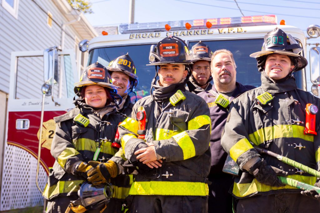 Five firefighters in full gear stand in front of a fire truck on a sunny day, posing and smiling for the camera. The fire truck behind them displays "Broad Channel V.F.D." on its sign.
