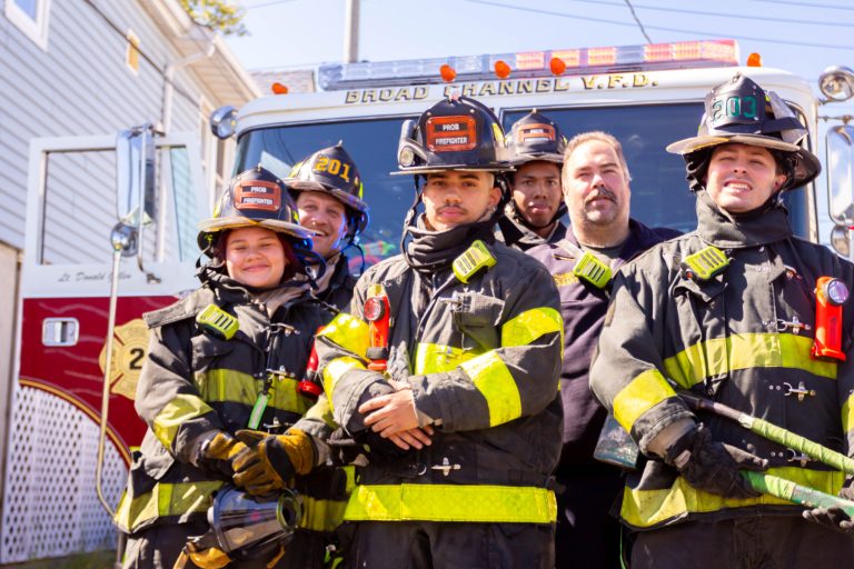 Five firefighters in full gear stand in front of a fire truck on a sunny day, posing and smiling for the camera. The fire truck behind them displays "Broad Channel V.F.D." on its sign.