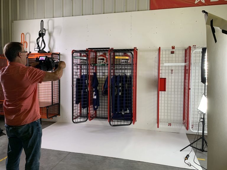 A man in a red shirt photographs red metal sports lockers holding helmets and uniforms in a studio with professional lighting equipment set up around him.
