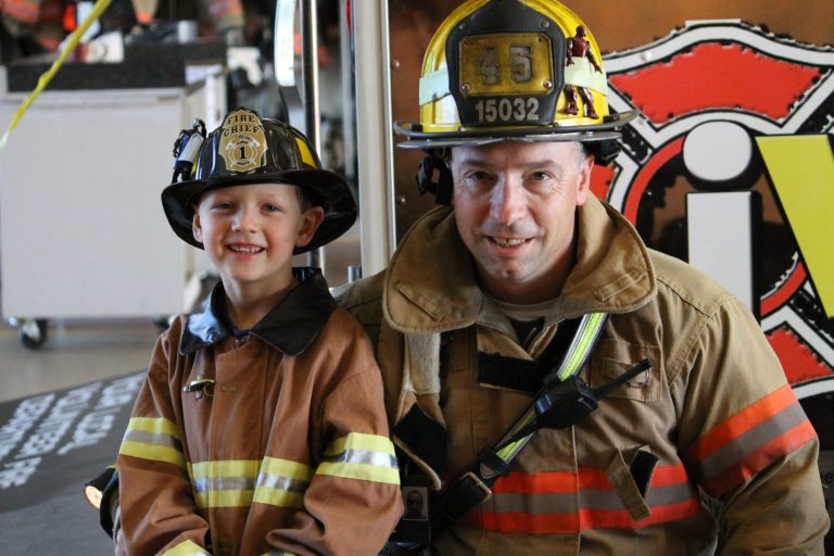 A firefighter and a smiling child, both in firefighter uniforms and helmets, pose together indoors in front of a fire truck.