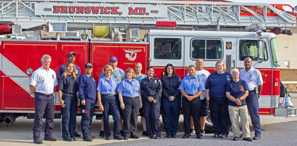 A group of firefighters and emergency personnel, both men and women, stand in uniform in front of a red Brunswick, MD fire truck, posing for a group photo outside on a sunny day.