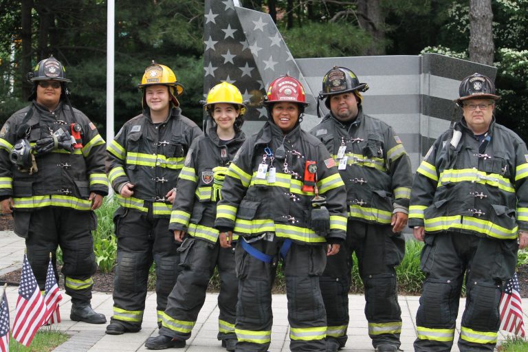 Six firefighters wearing full gear stand together and smile in front of a memorial with an American flag design. Small American flags border the walkway, and trees are visible in the background.
