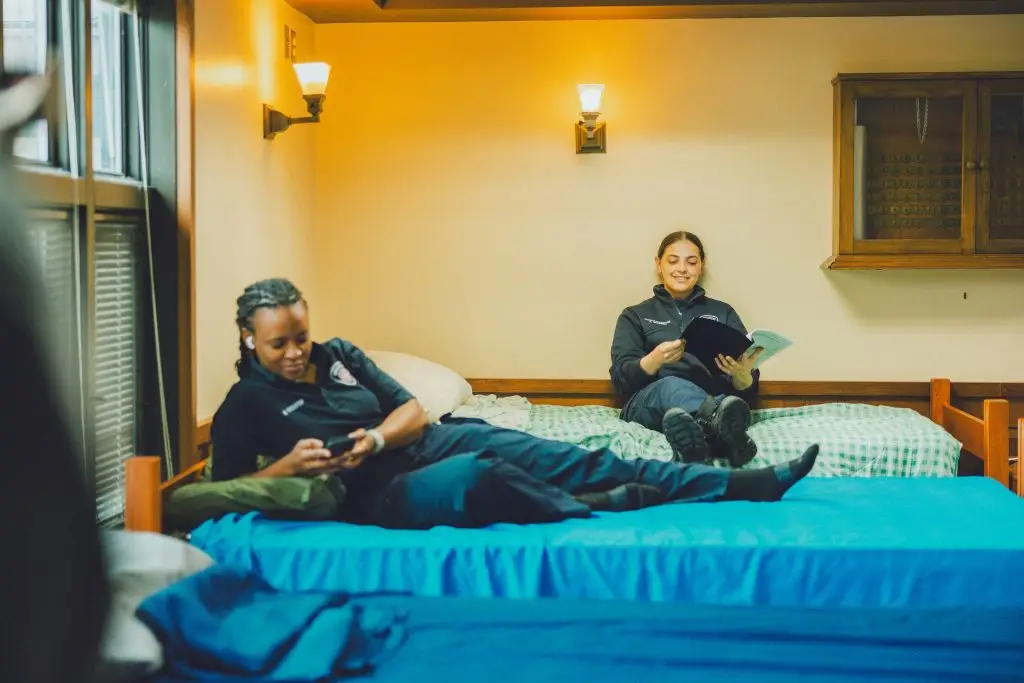 Two women in uniform relax in a dorm room; one is lying on a bed using her phone, while the other sits on a different bed smiling and reading a book. The room has soft lighting and neatly made beds.