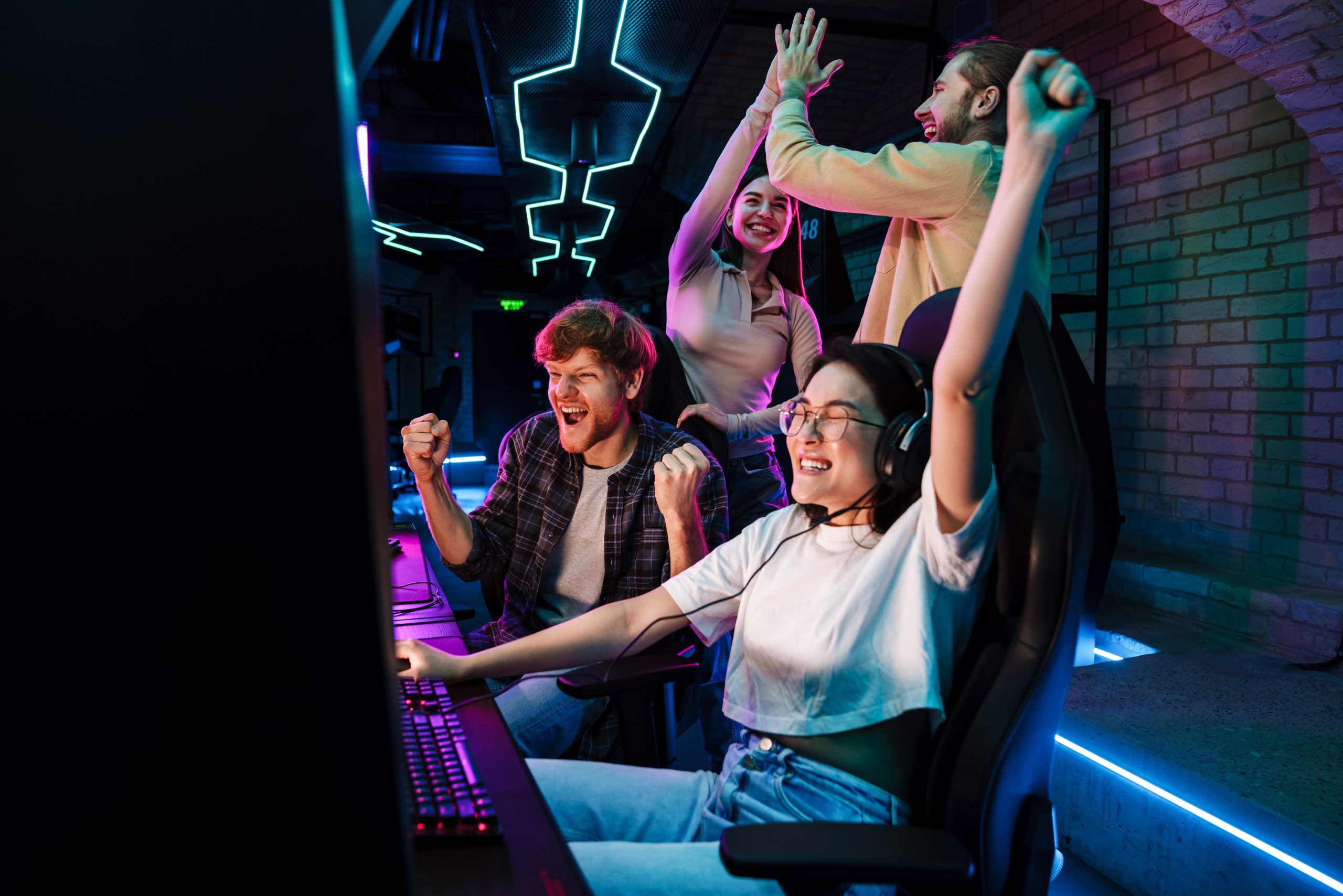 Four young adults celebrate at a gaming setup under neon lights; two are seated at computers cheering, while two others stand behind them giving high-fives and smiling.
