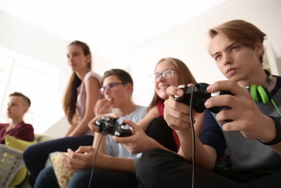A group of five teenagers sits together indoors; two in the front are playing video games with controllers, while the others watch. The atmosphere appears focused and relaxed.