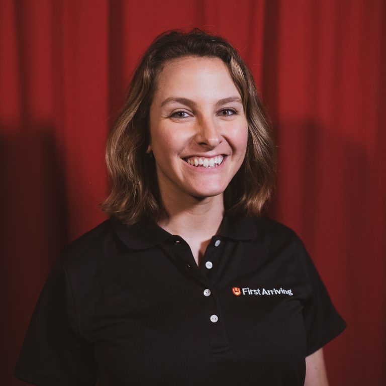 A woman with shoulder-length brown hair smiles while standing in front of a red curtain, wearing a black polo shirt with the "First Arriving" logo on it.