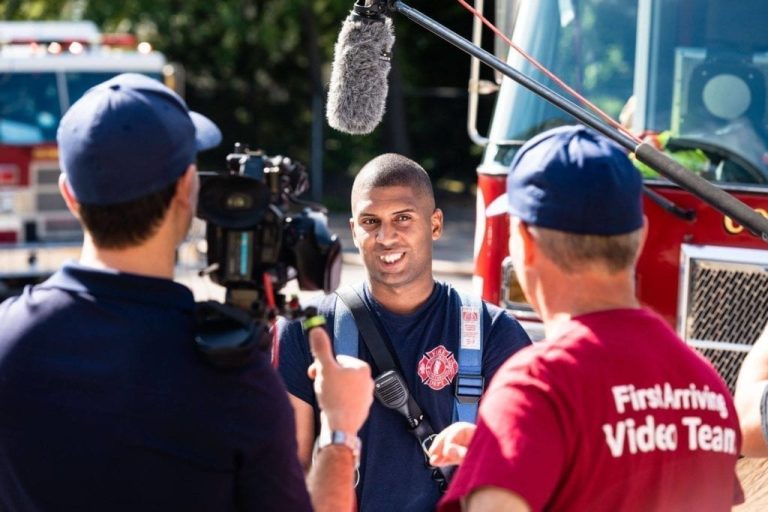 A firefighter stands in front of a fire truck, being interviewed on camera by a video crew. One crew member holds a microphone above the firefighter, while another wears a shirt labeled "FirstArriving Video Team.