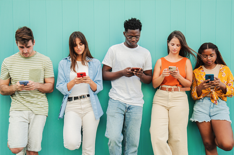 Five young adults stand against a turquoise wall, all looking down at their smartphones. They are casually dressed in light, summery clothes and appear focused on their devices.