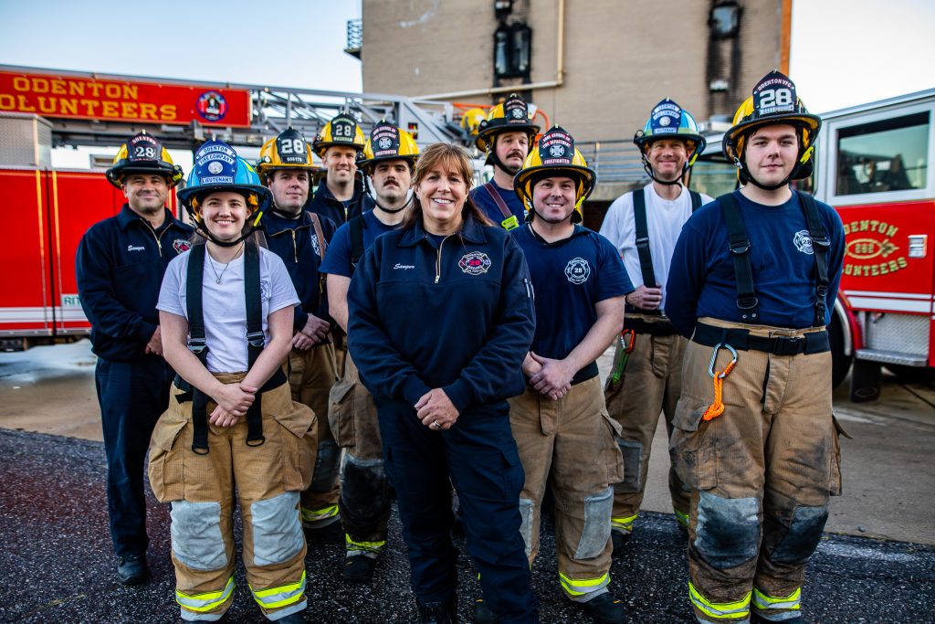 A group of firefighters in uniform and gear stand together outside, smiling at the camera. A fire truck labeled "Odenton Volunteers" is visible in the background.