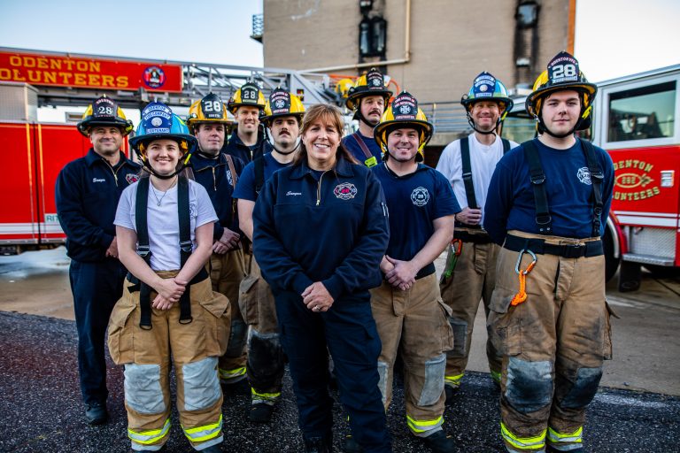 A group of firefighters in uniform and gear stand together outside, smiling at the camera. A fire truck labeled "Odenton Volunteers" is visible in the background.