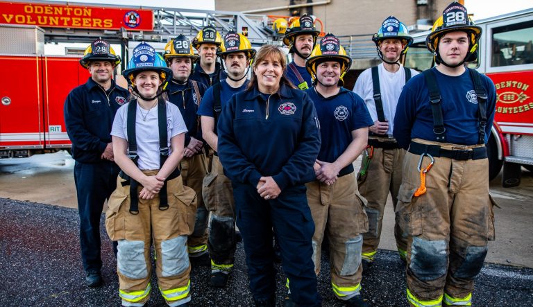 A group of firefighters in uniform and helmets stand smiling together in front of a red fire truck labeled "Odenton Volunteers" on a sunny day.