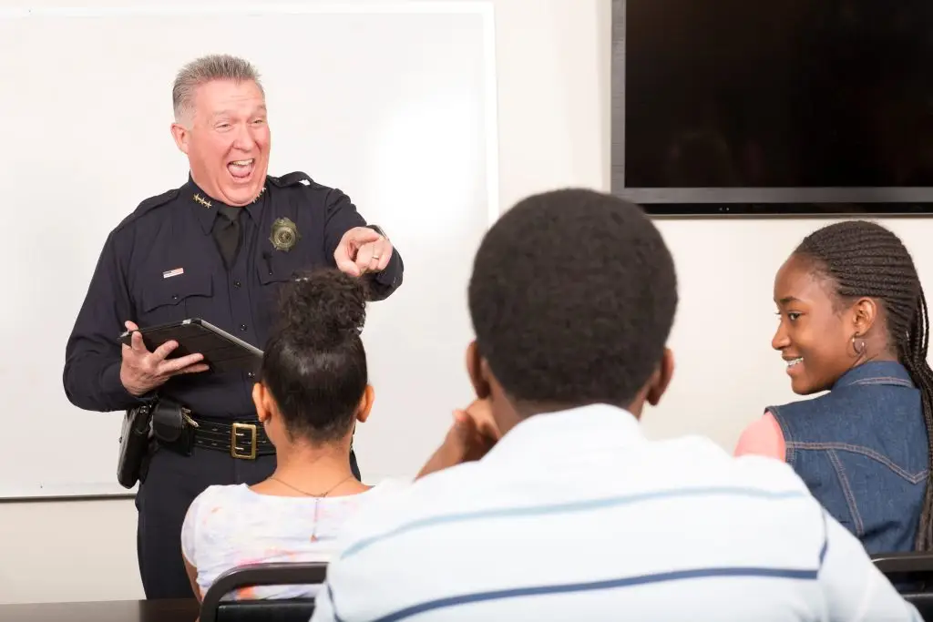 A police officer stands at the front of a classroom, smiling and pointing while holding a clipboard, as three students sit facing him and listen attentively.