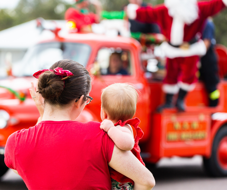 A woman holding a baby waves at a person dressed as Santa Claus on a festive red fire truck during a holiday parade. Both the woman and child wear red as they watch the celebration.