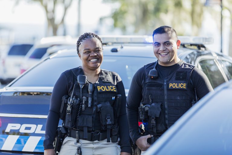 Two smiling police officers in black vests stand in front of a police car outdoors on a sunny day. The word "POLICE" is visible on their uniforms and on the vehicle behind them.