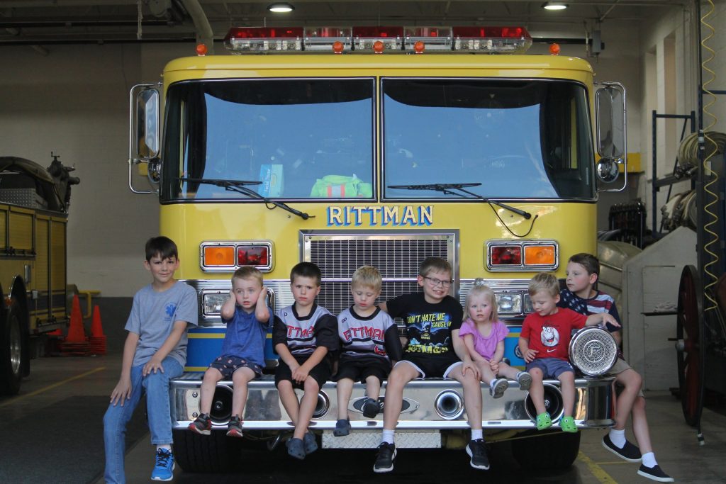 Eight children sit on the front bumper of a yellow fire truck labeled "Rittman" inside a fire station, smiling and posing for a photo.