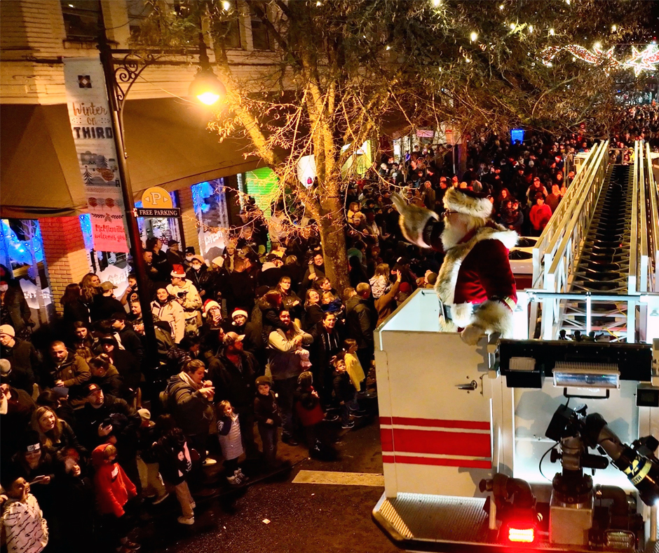 Santa Claus waves to a large crowd from the top of a fire truck during a nighttime holiday parade, with festive lights overhead and onlookers gathered along the street.