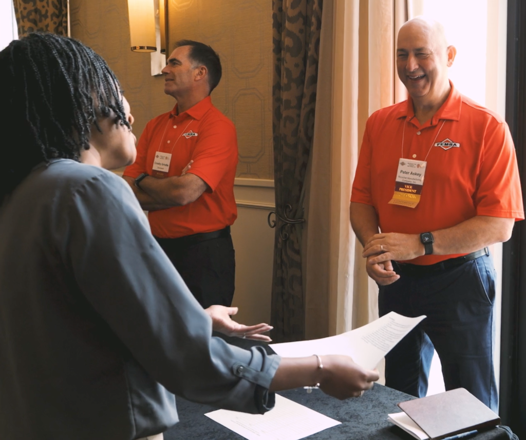 A woman hands papers to a smiling man wearing a red shirt and conference badge, while another man in a similar outfit stands nearby with arms crossed, inside a well-lit room.