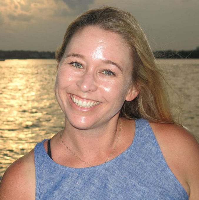 A woman with blonde hair and a blue sleeveless top smiles at the camera, standing outdoors near water during sunset, embodying the positive impact of public safety marketing in her community.