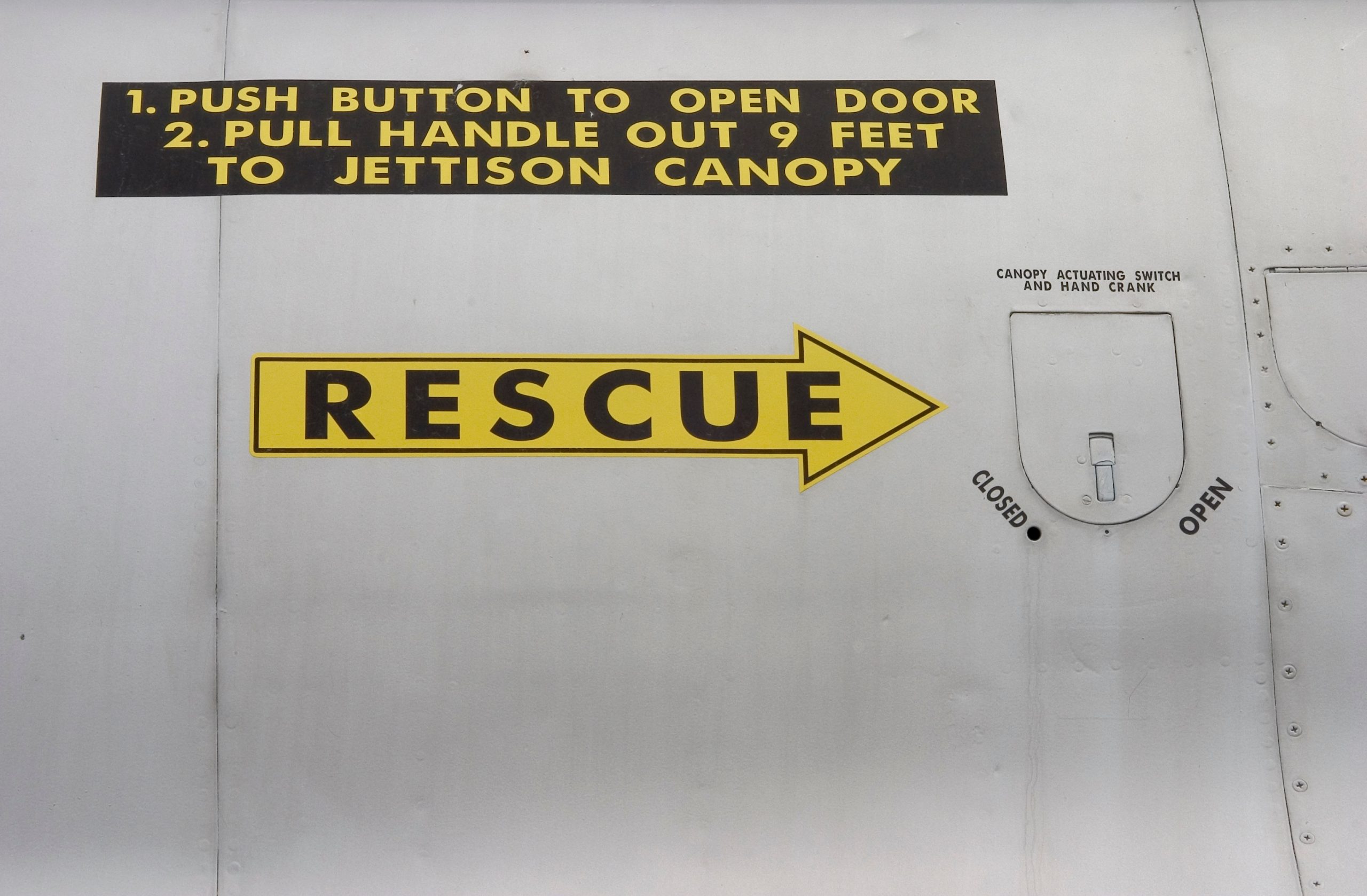 Yellow "RESCUE" sign with an arrow on the side of an aircraft, with instructions above to push a button and pull a handle to jettison the canopy. A labeled canopy actuating switch is also visible.