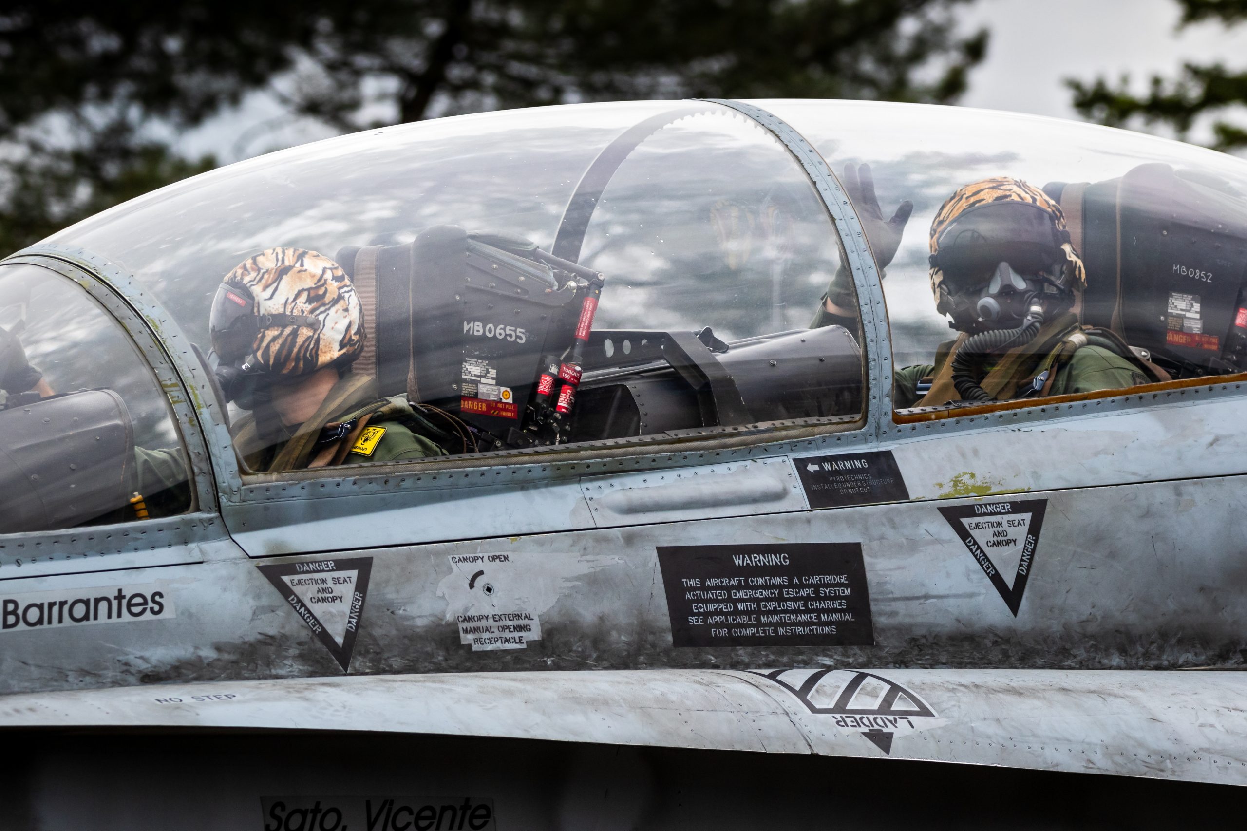 Two pilots wearing helmets and oxygen masks sit inside the cockpit of a military jet, with warning labels visible on the aircraft’s exterior and trees blurred in the background.