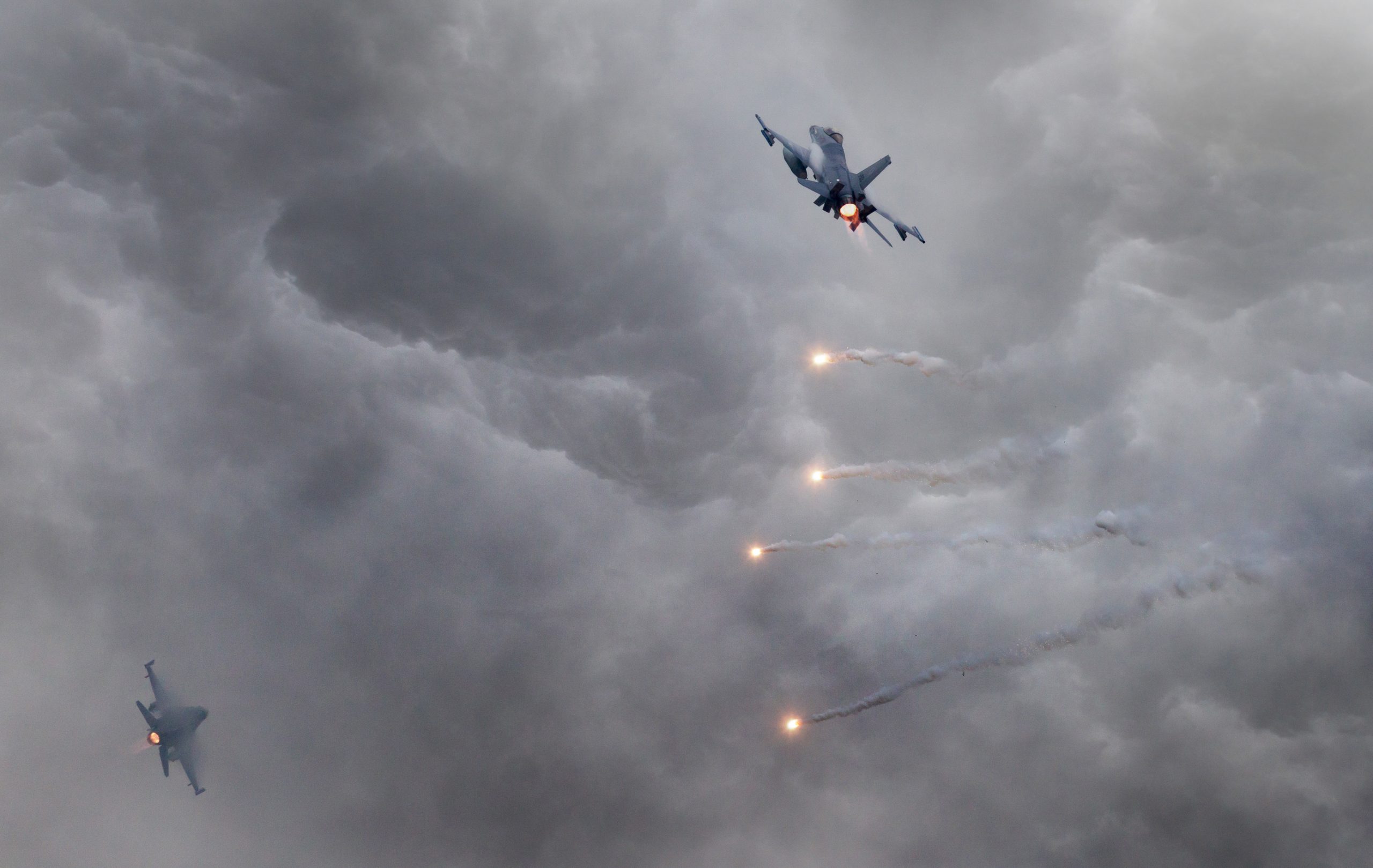 Two fighter jets fly through dramatic, cloudy skies with one jet releasing bright flares, leaving trails of smoke behind as they maneuver in the turbulent atmosphere.