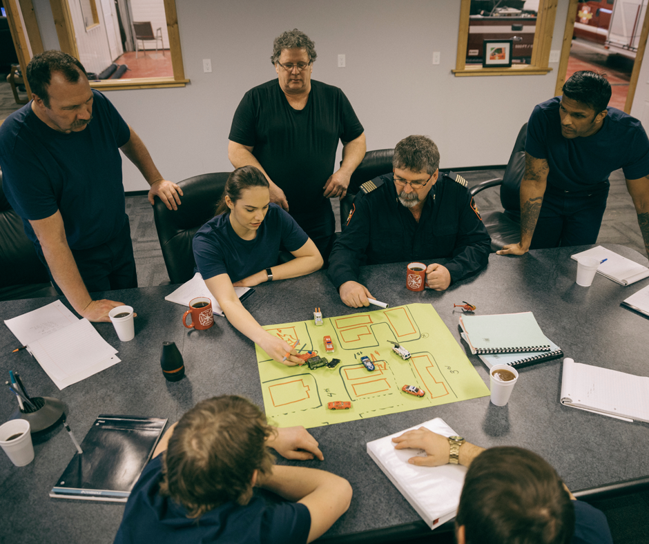Six people sit and stand around a table, discussing a tabletop layout with toy vehicles and a map. Notebooks, papers, and coffee cups are on the table, suggesting a planning or strategy meeting.